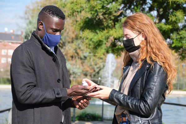 African black man and redhead caucasian woman wearing protective face masks using alcoholic gel in a park. Young multiracial couple during coronavirus pandemic.