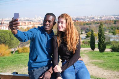 Afrikalı siyah bir adam ve kızıl saçlı bir kadın parkta gülüyor ve akıllı telefonlarıyla selfie çekiyorlar. Genç çok ırklı çift portresi.