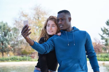 Afrikalı siyah bir adam ve kızıl saçlı bir kadın parkta gülüyor ve akıllı telefonlarıyla selfie çekiyorlar. Genç çok ırklı çift portresi.