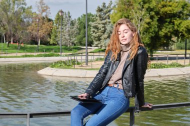 Kızıl saçlı kadın, parkta kitap okuyor ya da tablet veya e-kitapta çalışıyor. Dijital göçebe girişimci konsepti.