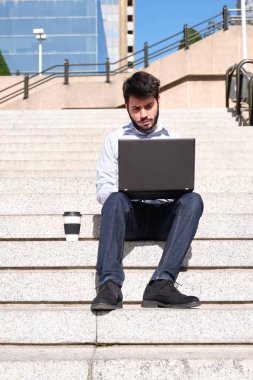 Genç iş adamı dizüstü bilgisayarını kullanıyor, elinde kahve fincanı, merdivenlerde oturuyor. Bir şehrin kentsel caddesinde iş adamı.