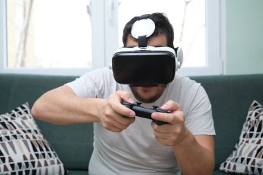 Sanal gerçeklik kulaklığıyla video oyunları oynayan genç bir adam ve kanepede oturan bir oyun platformu. Evde eğleniyorum..