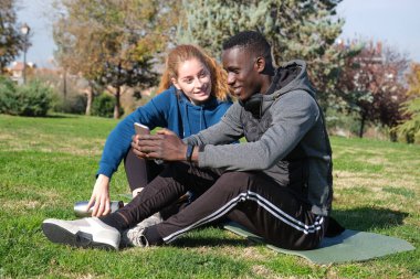 Afrikalı siyah bir adam ve kızıl saçlı bir kadın parkta hasırla egzersiz yaptıktan sonra akıllı telefonu kontrol ediyorlar. Spor ve spor açık havada.