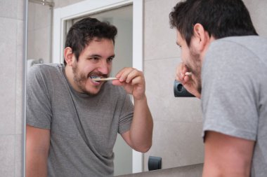 Genç latin adam sabah banyoda aynanın önünde dişlerini fırçalıyor. Çevre dostu bambu diş fırçası..