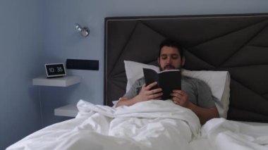 Genç latin adam sabahın geç saatlerinde yatakta kitap okuyor. Arka planda dijital saat var. 4k video.