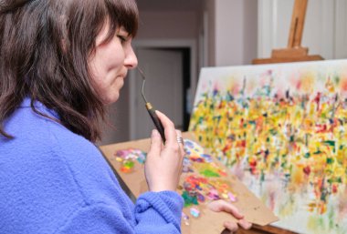 Genç bir kadın evde yağlı boyalarla tuval üzerinde düşünüyor ve resim yapıyor. Resim sehpasında sanatçı çizimi.