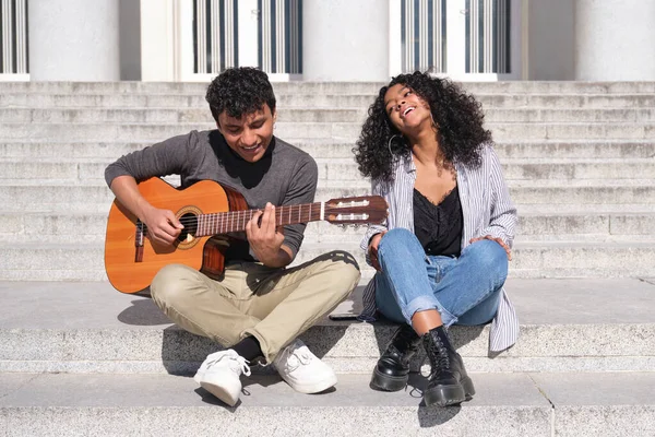 A young latin couple playing the guitar and singing sitting on stairs. Millennial generation.