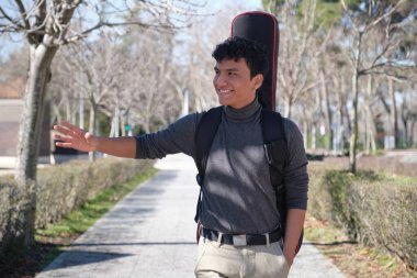 Latin genç bir adam gitar kılıfında gitar taşıyor, gülümsüyor ve şehirde birine el sallıyor. Üniversite kampüsü.