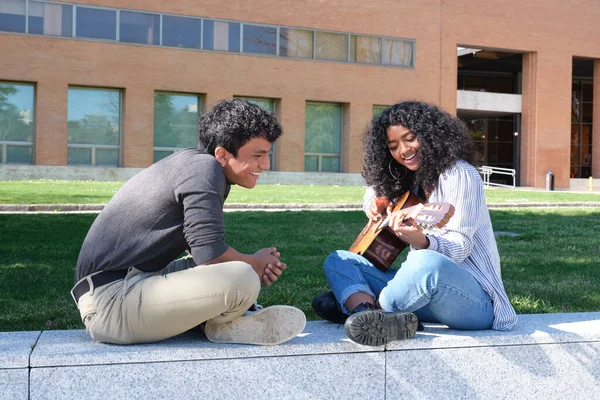 Young latin man teaching how to play the guitar to a young latin woman at the university campus. University life, millennial generation.