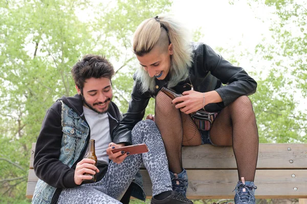 若いパンクカップルはベンチに座って 公園でスマートフォンをチェック笑ってビールを飲んでいます ロック ロールライフスタイル — ストック写真