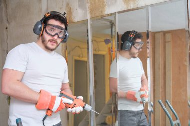 İnşaat işçisi elinde matkapla şantiyedeki kameraya bakıyor. Güvenlik eldiveni, gözlük ve kulaklık takan inşaat işçisi..