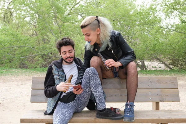 若いパンクカップルはベンチに座って公園でスマートフォンをチェックしてビールを飲んでいます ロック ロールライフスタイル — ストック写真