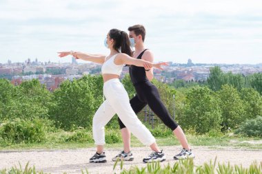 Erkek eğitmen, savaşçı yoga pozuyla parkta koruyucu maskeler takan genç bir kadına yardım ediyor. Salgın sırasında dışarıda spor yapmak.