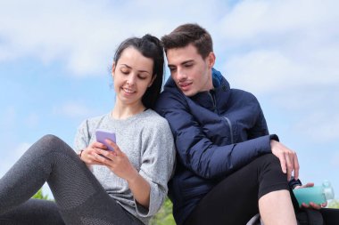 Parkta otururken akıllı telefonu kontrol eden mutlu genç çift. Fitness teknolojisi kavramı.