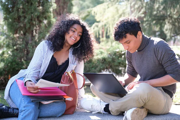 İki Latin öğrenci dışarıda bir bankta oturup ders çalışırken gülümsüyor. Kampüste üniversite hayatı.