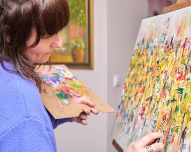 Evde yağlı boyalarla tuvale resim yaparken gülümseyen genç bir kadın. Resim sehpasında sanatçı çizimi.