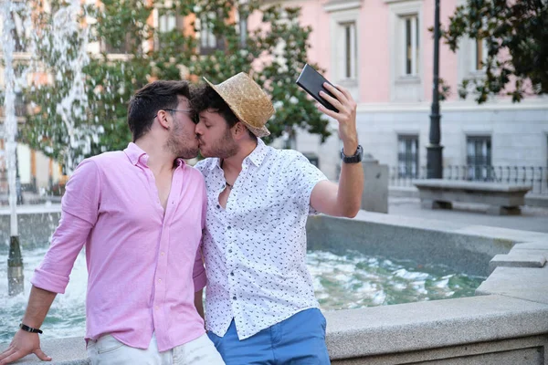 Egy meleg turista pár csókolózik és szelfit készít a szökőkútnál egy városban.. — Stock Fotó