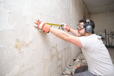 İnşaatçı inşaat alanında bir mezura kullanıyor. Güvenlik eldiveni, gözlük ve kulaklık takan inşaat işçisi..