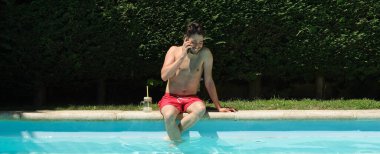 Yüzme havuzunun kenarında oturmuş akıllı telefonuyla konuşan genç bir adamın panoramik görüntüsü. Yaz konsepti.