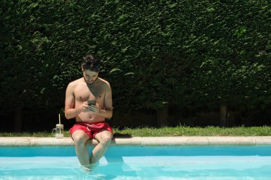 Havuzun kenarında oturan akıllı telefonuyla dijital göçebe çalışıyor. Yaz konsepti.