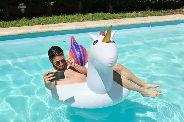 一个戴着太阳镜的年轻人在游泳池里戴着一只独角兽充气环自拍 夏季概念 — 图库照片