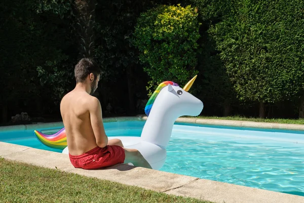年轻人坐在游泳池边 手里拿着一个大独角兽充气环 夏季概念 — 图库照片