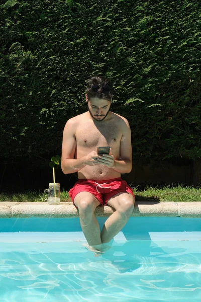 Giovanotto Che Usa Suo Smartphone Seduto Sul Bordo Della Piscina — Foto Stock