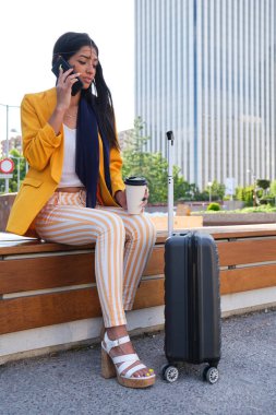 Genç Hintli iş kadını elinde kahve ve bavuluyla telefonla konuşuyor..