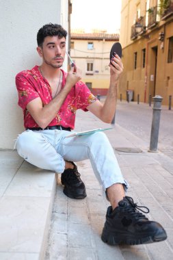 Uzun takma tırnaklı beyaz bir adam caddede aynanın önüne kırmızı far sürüyor..