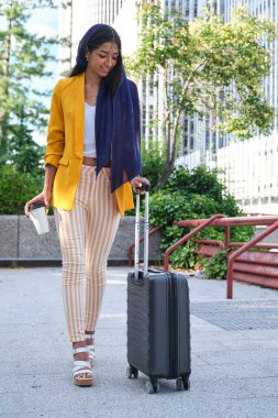 Sarı takım elbiseli genç Hintli iş kadını elinde kahve ve açık havada bir valizle yürüyüp duruyor..