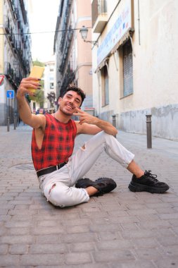 Uzun takma tırnaklı beyaz bir adam yerde oturan akıllı telefonuyla selfie çekiyor..
