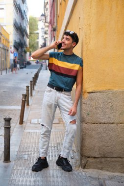 Uzun takma tırnakları olan, akıllı telefonda gülümseyen ve konuşan beyaz bir adam, sarı bir duvarın önünde duruyor..