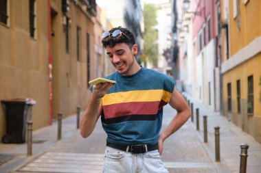 Uzun takma tırnaklı, beyaz bir genç adam, gülümsüyor ve bir şehir caddesinde akıllı telefonuyla bir ses kaydı yapıyor..