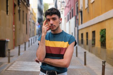 Uzun takma tırnaklı, kablosuz kulaklıklı, caddeye bakan genç, beyaz bir adam..