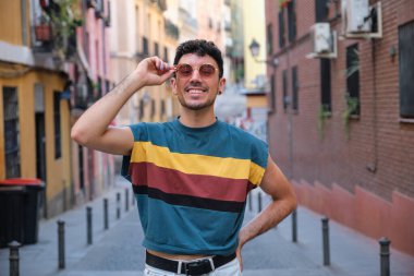 Uzun takma tırnaklı, güneş gözlüğüyle poz veren beyaz bir adam sokağa bakıyor..