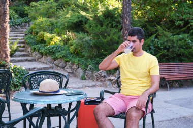 Turist dinleniyor, serinliyor ve yaz tatili terasında bavulu ve şapkasıyla oturuyor..