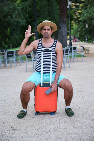 Tourist man gesturing pretending to have gun sitting on wheeled suitcase in a park.