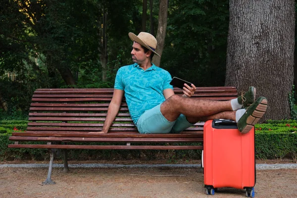 Tourist man using the smartphone sitting on a bench with his feet on the suitcase and a straw hat.