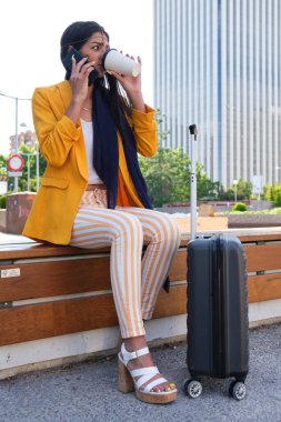 Sarı takım elbiseli genç Hintli iş kadını akıllı telefonla konuşuyor, açık havada duran bavuluyla kahve içiyor..