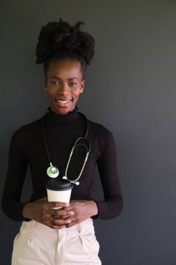 Steteskoplu gülümseyen Afrikalı kadın doktor portresi ve siyah arka planda bir fincan kahve. Stüdyo çekimi.