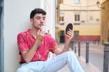 Uzun takma tırnaklı beyaz bir adam caddede aynanın önüne kırmızı ruj sürüyor..