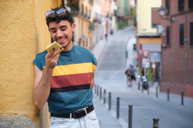 Uzun takma tırnakları olan beyaz bir genç adam, gülümsüyor ve sarı bir duvarın karşısında duran akıllı telefonuyla bir ses kaydediyor..