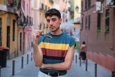 Uzun takma tırnaklı, güneş gözlüğüyle poz veren beyaz bir adam sokağa bakıyor..