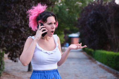 Pembe saçlı, beyaz, genç bir kadın parktaki akıllı telefondan konuşuyor..