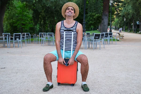 Tourist man laughing sitting on wheeled suitcase in a park.