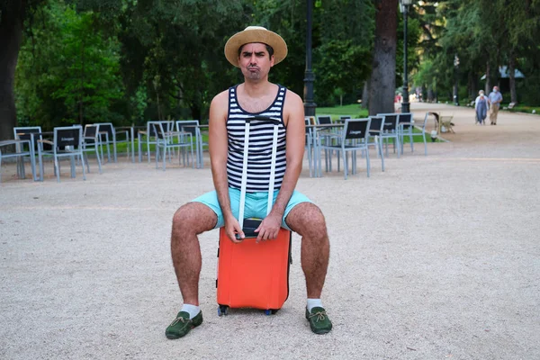 Tourist man with a suspicious face sitting on wheeled suitcase in a park.