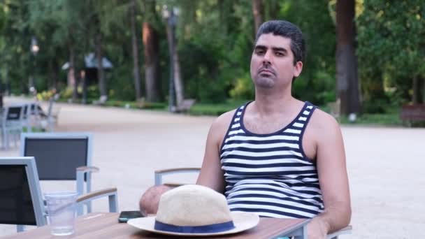 Турист бросает шляпу в камеру и улыбается сидя на террасе бара. — стоковое видео