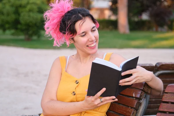 Νεαρή λευκή γυναίκα με ροζ μαλλιά κάθεται σε ένα παγκάκι και διαβάζει ένα βιβλίο. — Φωτογραφία Αρχείου