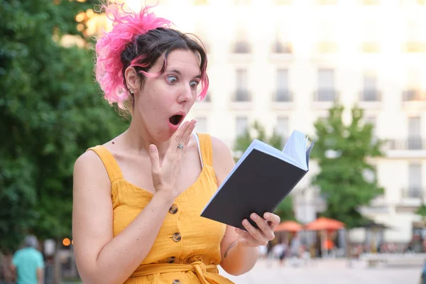 Νεαρή καυκάσια γυναίκα με ροζ μαλλιά διαβάζει ένα βιβλίο έκπληκτη. — Φωτογραφία Αρχείου