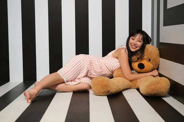 Junge asiatische Frau umarmt und legt sich über einen Teddybär auf den gestreiften Zimmerboden. — Stockfoto